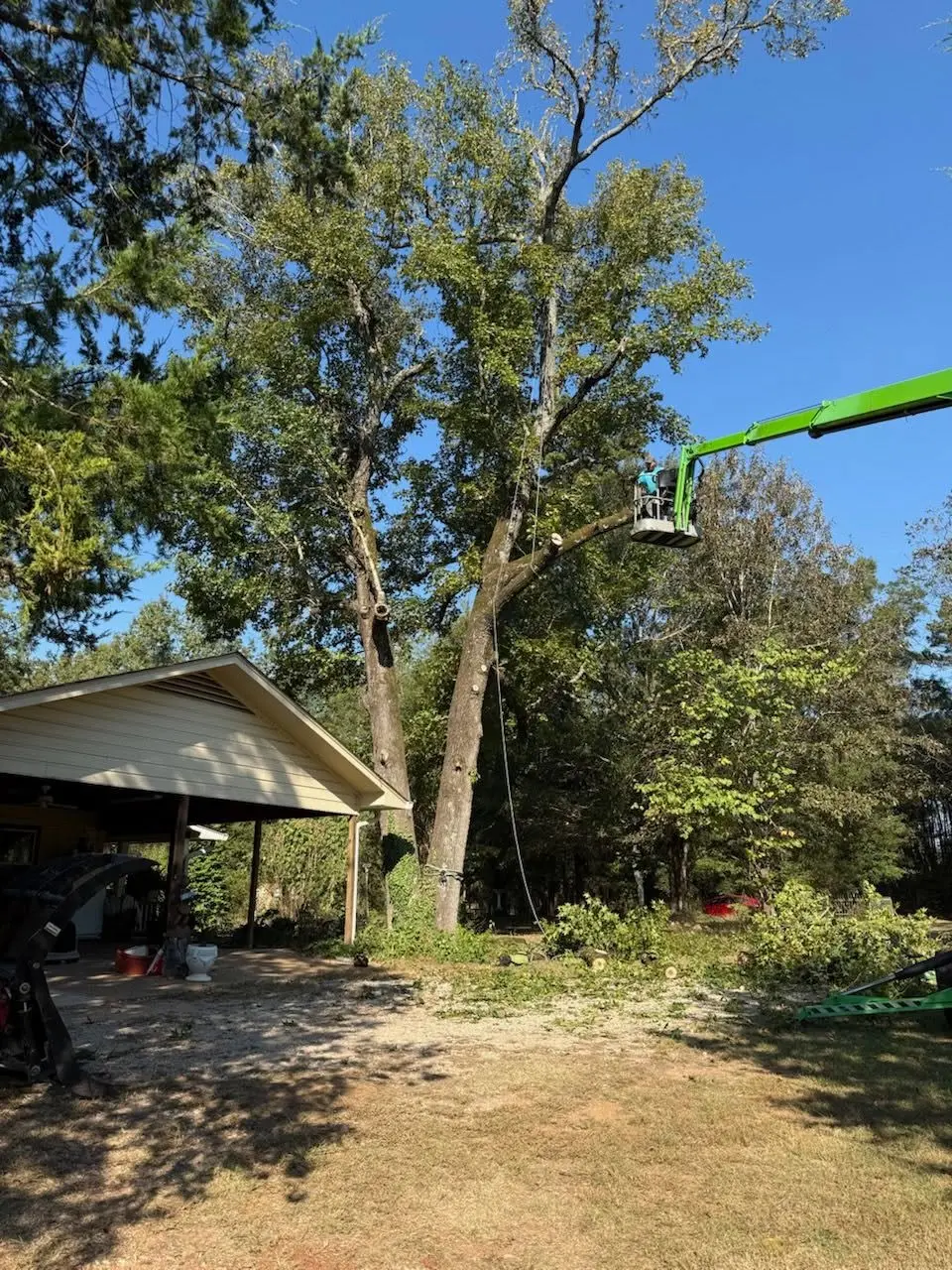 Boom lift cutting large limbs from a tree beside an open carport in Overton, Texas